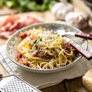 Spaghetti mit Prosciutto, Champignons und geriebenem Parmesan in einer gemusterten Schale, daneben eine Reibe und Petersilie