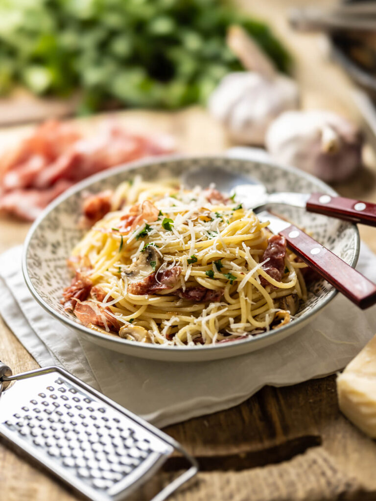 Spaghetti mit Prosciutto, Champignons und geriebenem Parmesan in einer gemusterten Schale, daneben eine Reibe und Petersilie