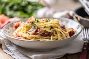 Teller mit Spaghetti, Prosciutto, Champignons und Petersilie, im Hintergrund frische Kräuter und Knoblauch