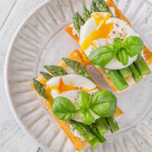 Zwei Ciabatta-Bruschette mit grünem Spargel, pochiertem Ei mit laufendem Dotter und frischem Basilikum auf hellem Teller