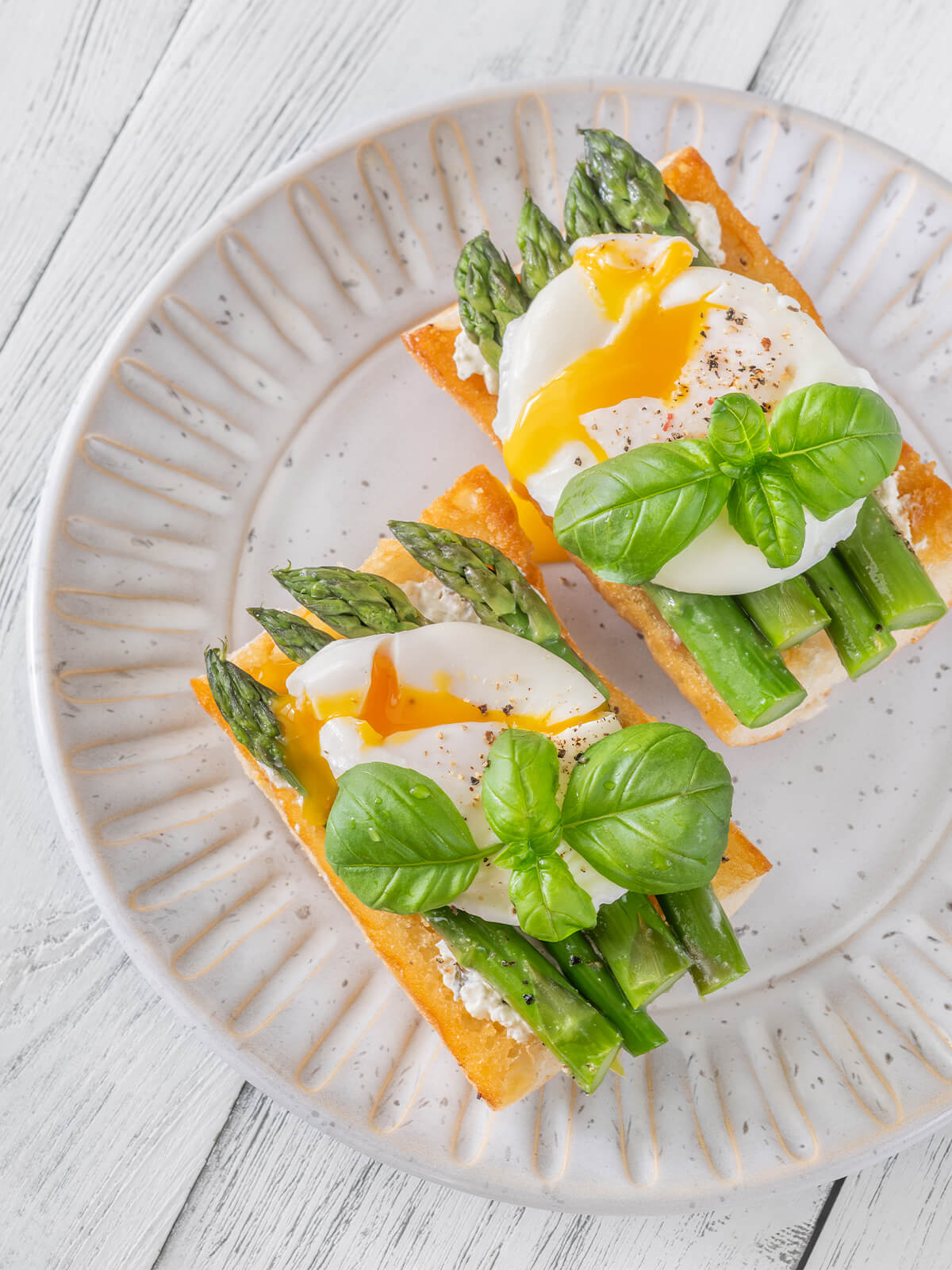 Zwei Ciabatta-Bruschette mit grünem Spargel, pochiertem Ei mit laufendem Dotter und frischem Basilikum auf hellem Teller