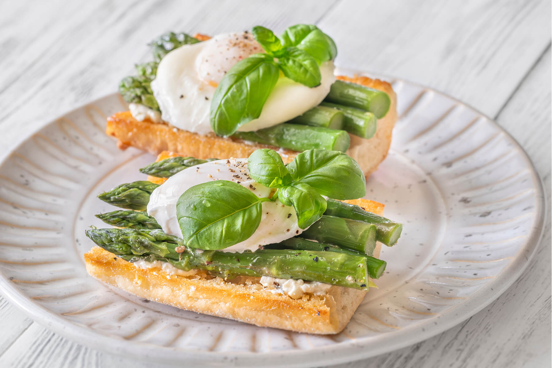Schräge Nahaufnahme von zwei Ciabatta-Bruschette mit grünem Spargel, pochiertem Ei und Basilikum auf hellem Teller