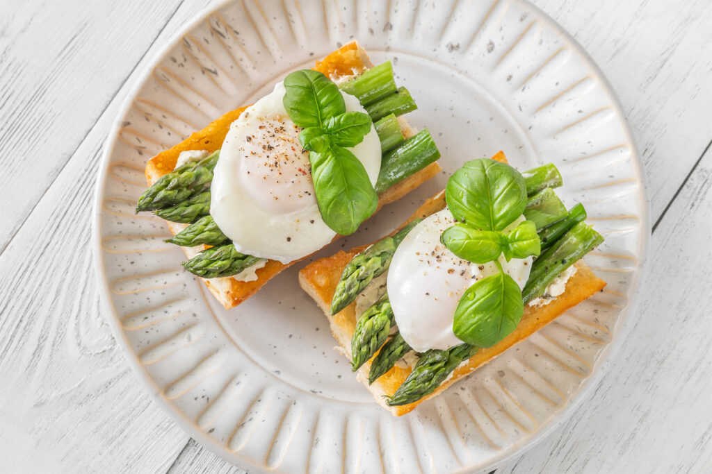 Zwei Ciabatta-Bruschette mit grünem Spargel, pochiertem Ei und Basilikum von oben auf hellem Teller