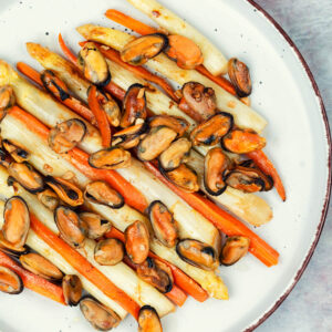 Warmer Spargelsalat mit ganzen weißen Spargelstangen, langen Karottenstiften und gebratenem Miesmuschelfleisch mit Knoblauchstücken auf einem hellen Teller