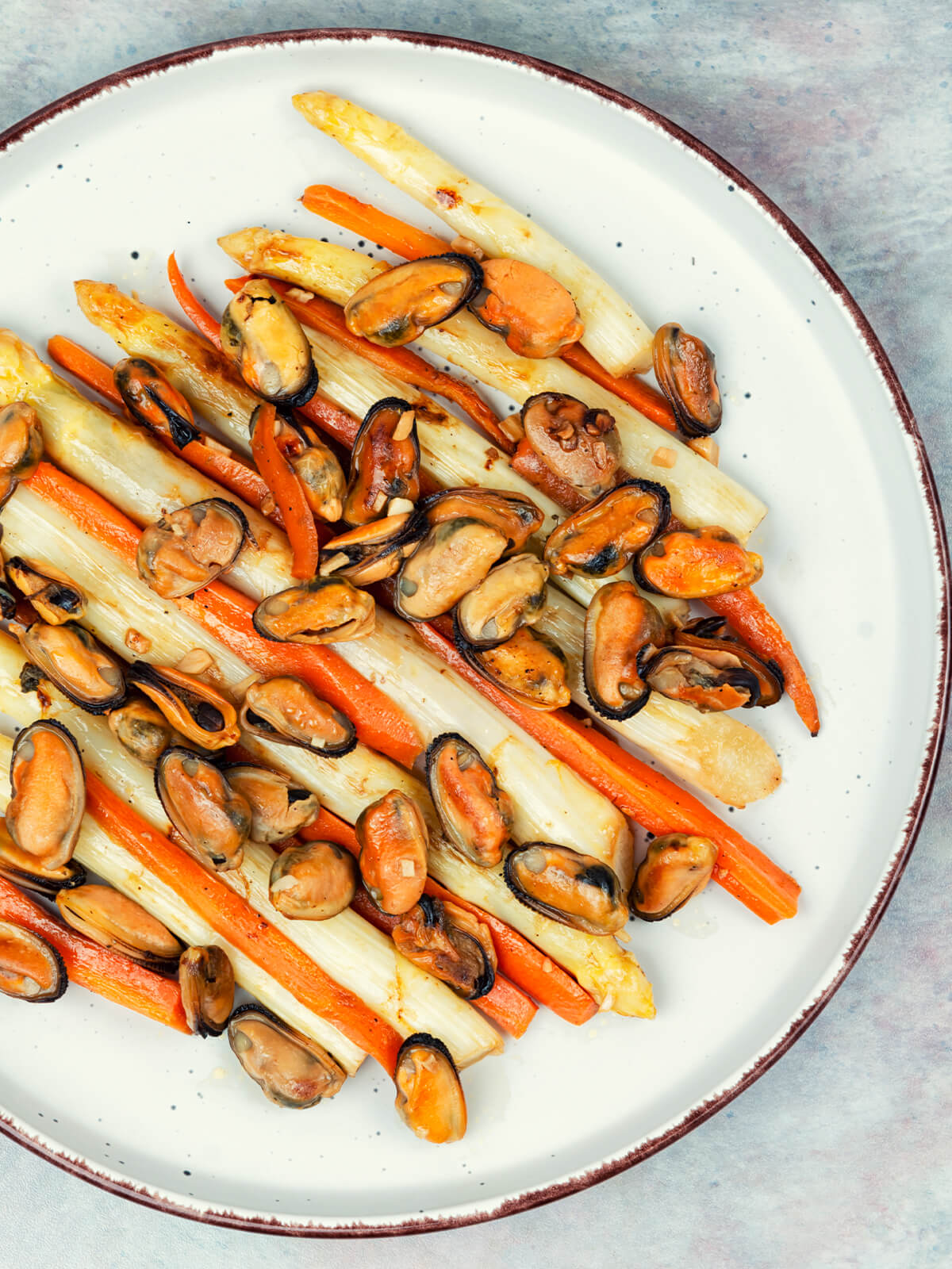 Warmer Spargelsalat mit ganzen weißen Spargelstangen, langen Karottenstiften und gebratenem Miesmuschelfleisch mit Knoblauchstücken auf einem hellen Teller