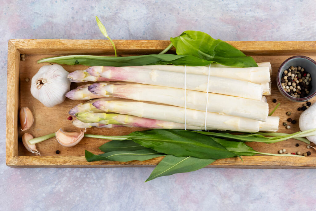 Bund weißer Spargel auf einem Holzbrett, daneben Knoblauchknollen und -zehen, eine Schale bunter Pfeffer und grüne Wildkräuterblätter