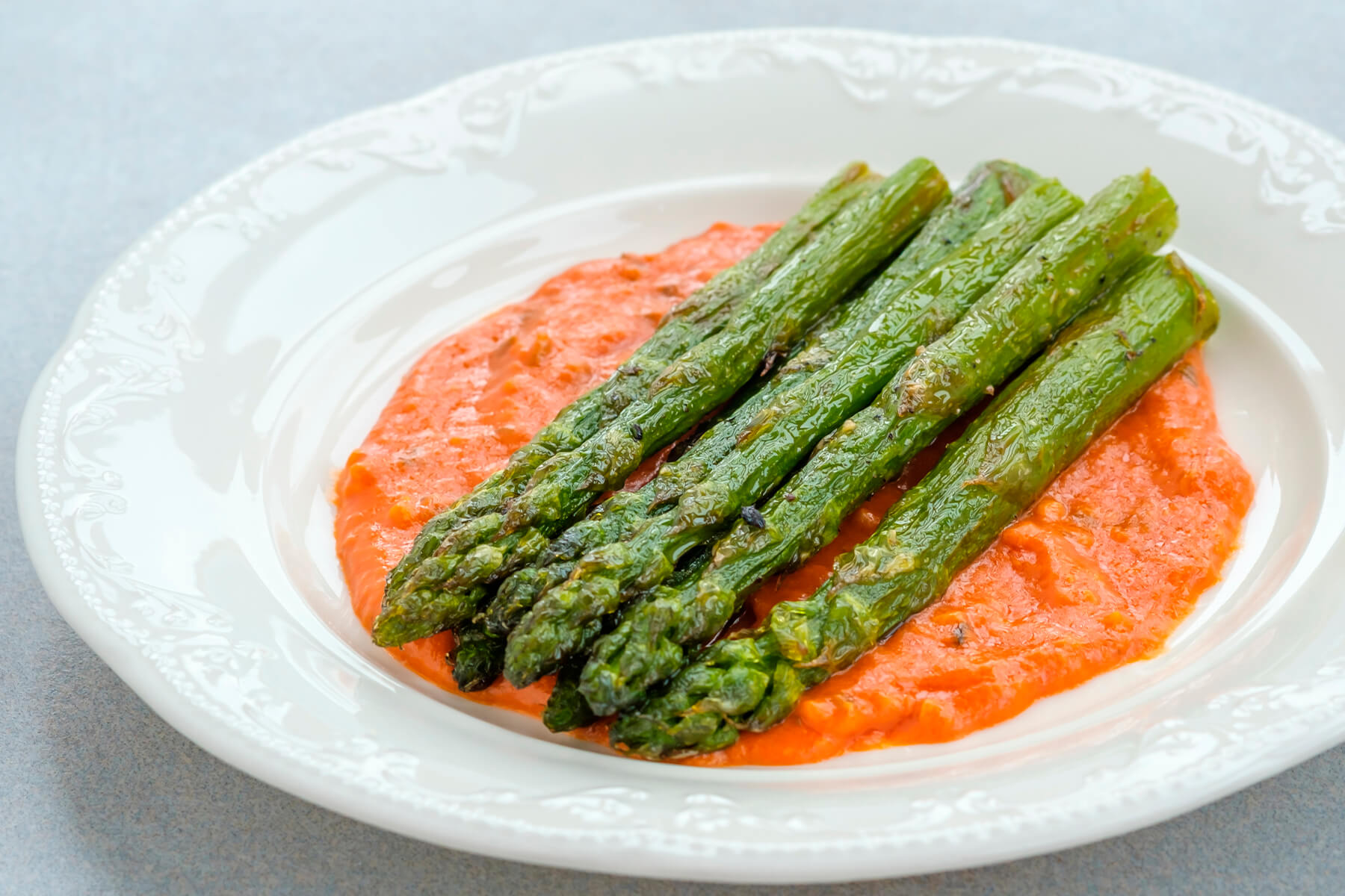 Gebratener grüner Spargel mit Röstaromen auf orangeroter Romesco-Sauce auf weißem Teller von der Seite