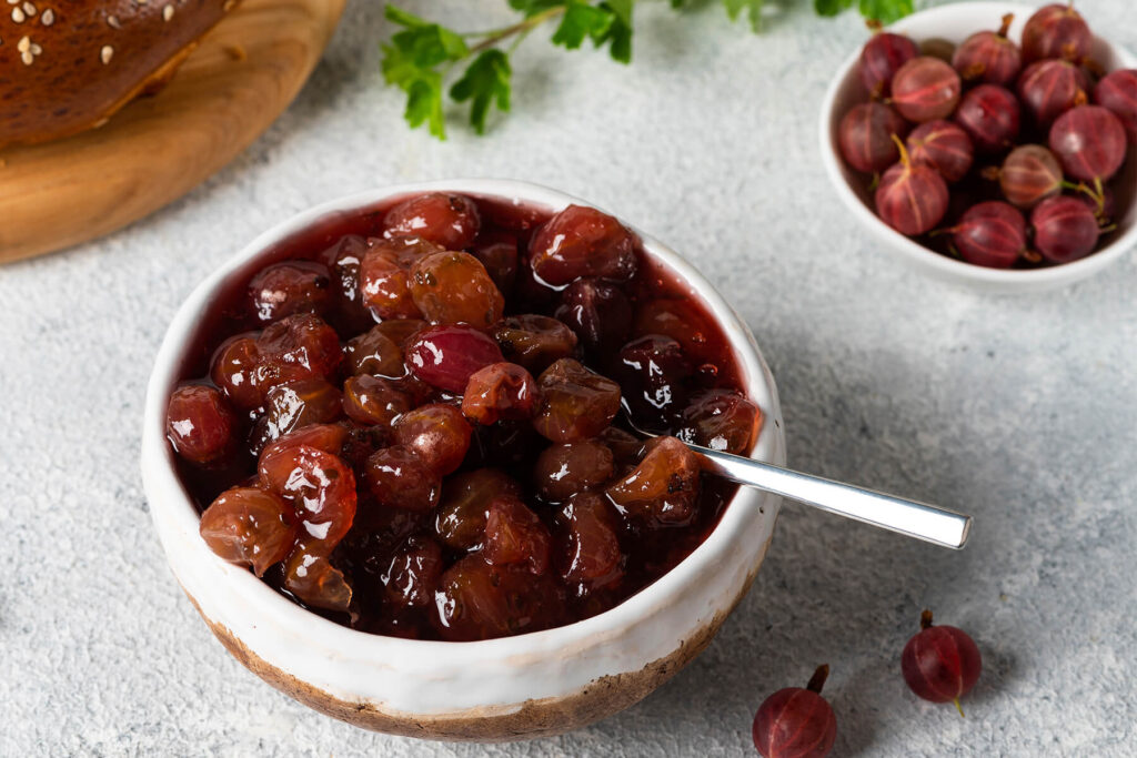Stachelbeer-Konfitüre mit ganzen Früchten in einer Schale, daneben frische Stachelbeeren und ein Sesambrötchen