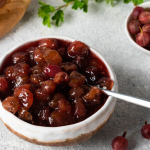 Stachelbeer-Konfitüre mit ganzen Früchten in einer Schale, daneben frische Stachelbeeren und ein Sesambrötchen