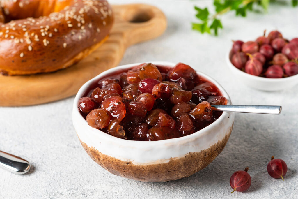 Fertige Stachelbeer-Konfitüre mit ganzen Beeren in einer Schale, dahinter ein Sesambrötchen