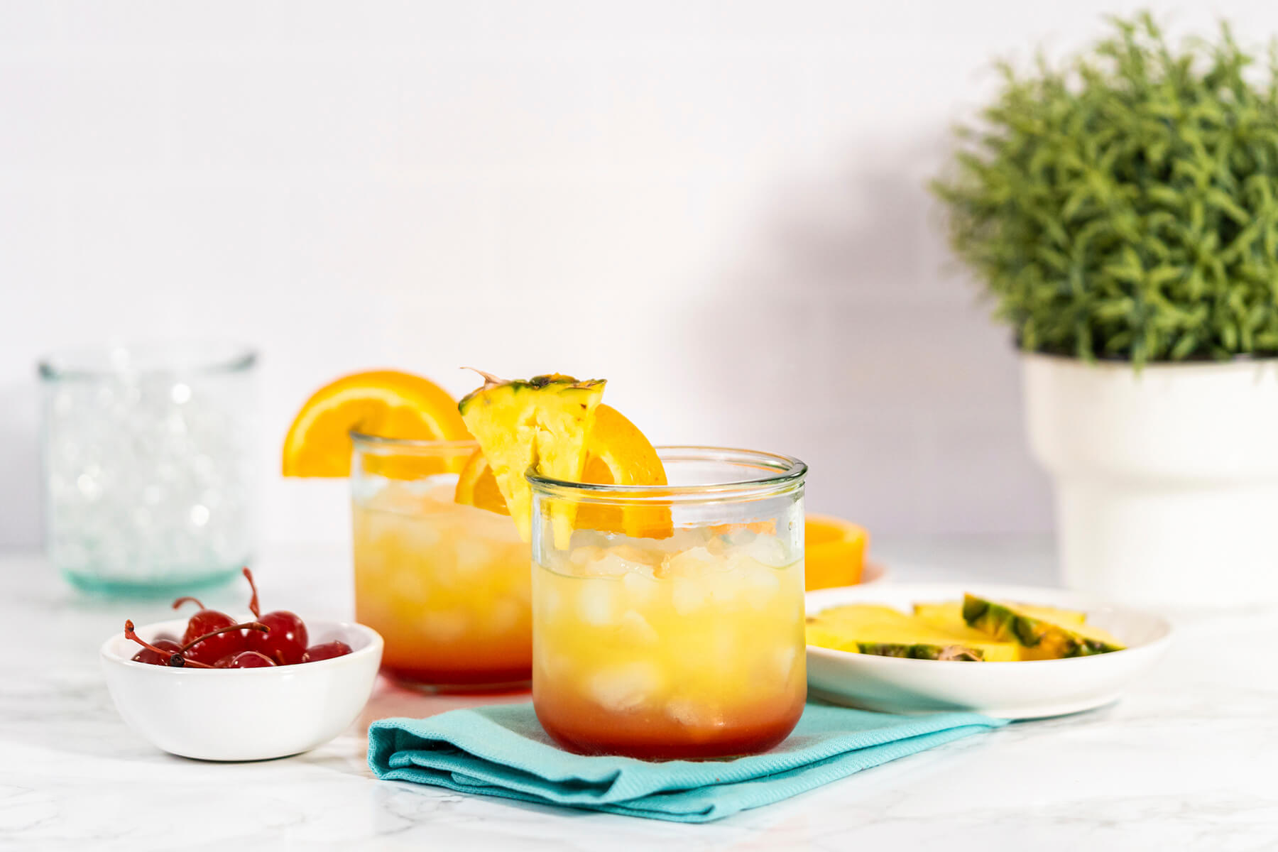 Zwei fertige Sunset-Cocktails mit Fruchtdeko nebeneinander, daneben Cocktailkirschen und Ananasstücke