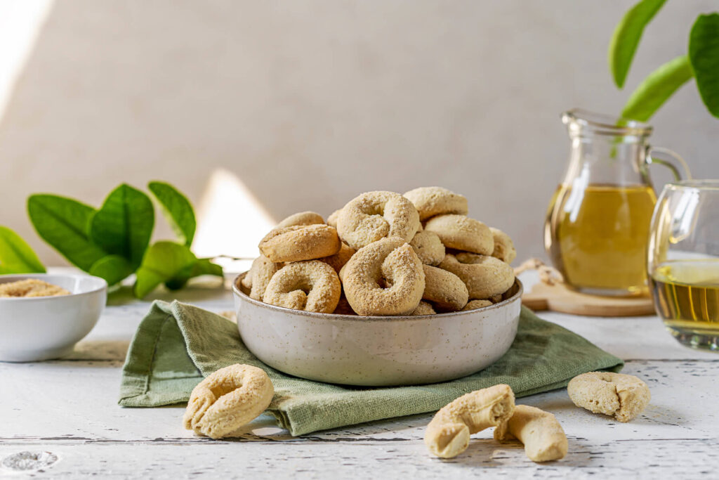 Süße Taralli dolci als kleine helle Kringel mit Zuckerkruste auf der Oberseite in einer Schale, dahinter ein Glas Weißwein