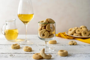 Fertige süße Taralli dolci mit heller Zuckerkruste auf der Oberseite in einem Stielglas, daneben ein Glas Weißwein