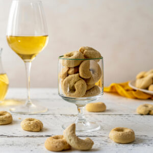 Fertige süße Taralli dolci mit heller Zuckerkruste auf der Oberseite in einem Stielglas, daneben ein Glas Weißwein