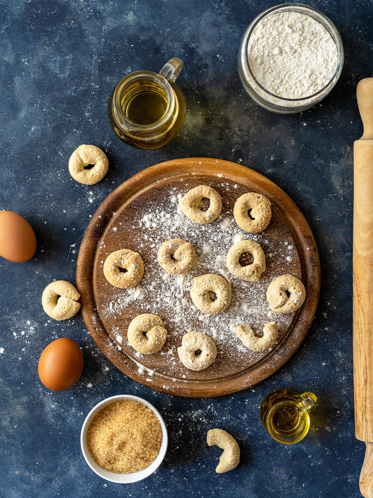Fertig gebackene süße Taralli dolci als kleine Kringel auf einem Holzbrett, daneben Eier, Mehl, Zucker und Olivenöl