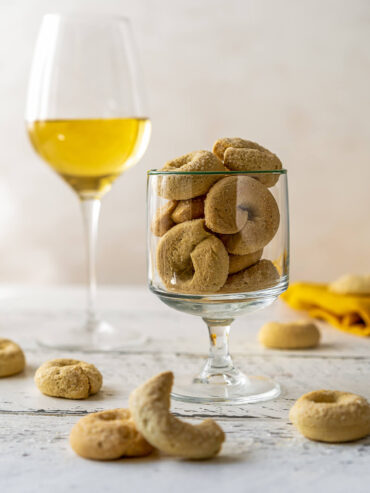Süße Taralli dolci mit heller Zuckerkruste auf der Oberseite in einem Stielglas, daneben ein Glas Weißwein – ein Weingebäck aus Apulien