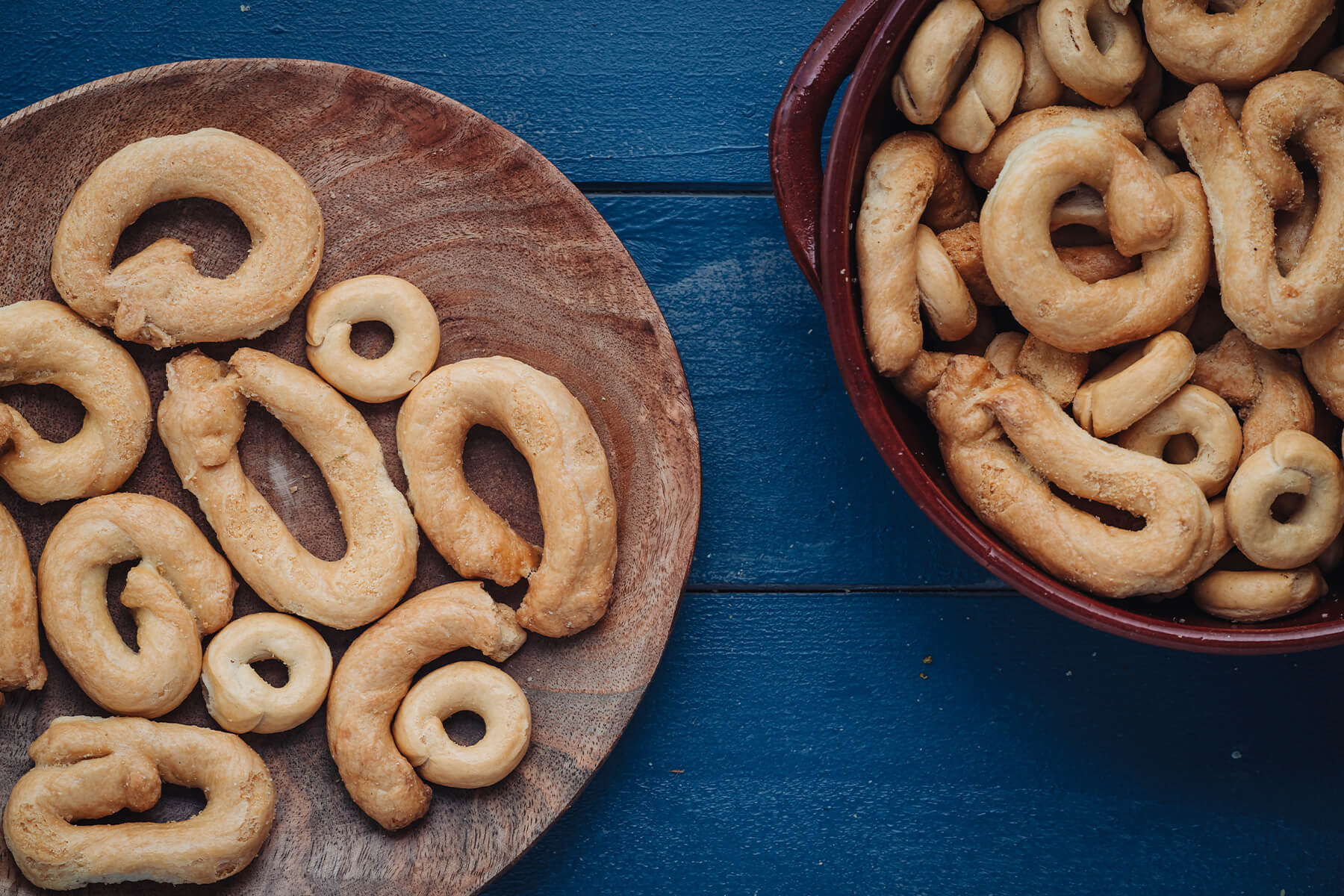 "Herzhafte Taralli Pugliesi von oben – goldbraune knusprige Kringel auf einem Holzteller und in einer Terrakotta-Schale auf blauem Tisch"