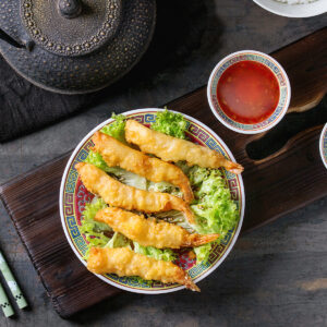 Fünf Tempura-Garnelen mit hell goldgelber, geschlossener Teighülle und teigfreien orange-bernsteinfarbenen Schwanzfächern auf krausem grünem Salat mit hellem Sesam, auf einem asiatischen Porzellanteller mit rotem Spiegel und buntem Mäanderrand; daneben Schälchen mit süßer Chilisauce und Sojasauce auf dunklem Holzbrett, dazu eine gusseiserne Teekanne und grüne Stäbchen