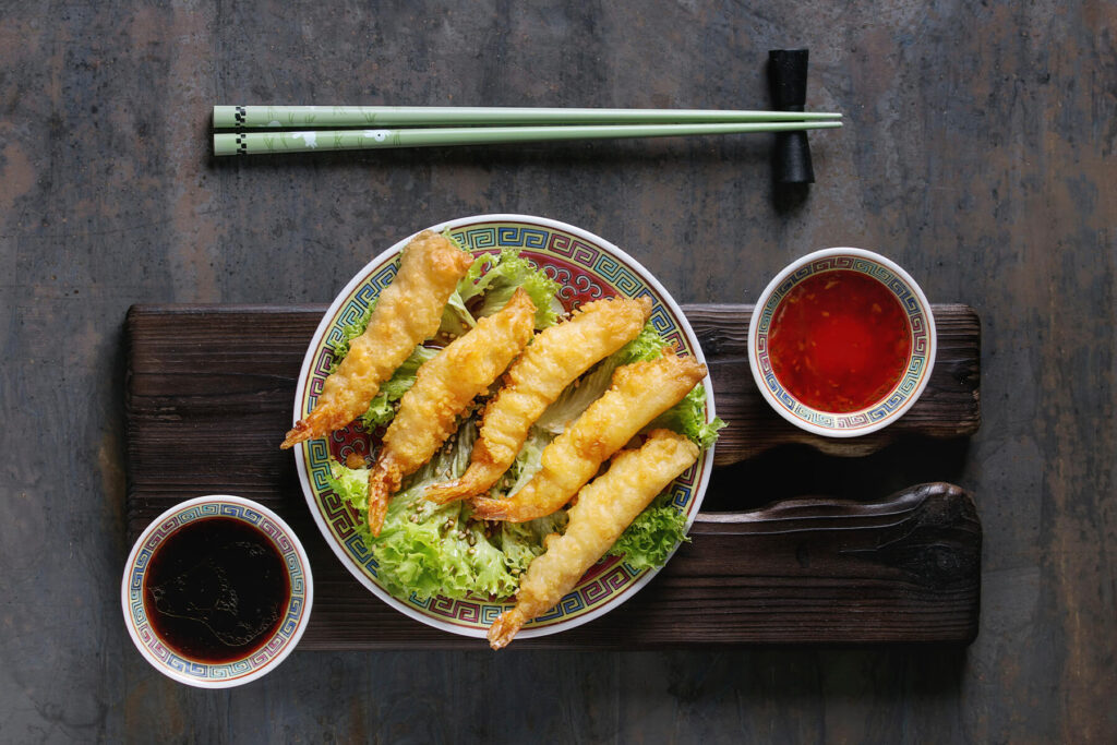 Angerichtete Tempura-Garnelen von oben: fünf Garnelen mit hell goldgelber, geschlossener Teighülle und teigfreien orange-bernsteinfarbenen Schwanzfächern, schräg überlappend auf krausem grünem Salat mit hellem Sesam, auf einem asiatischen Porzellanteller mit rotem Spiegel und buntem Mäanderrand auf dunklem Holzbrett, daneben Schälchen mit süßer Chilisauce und Sojasauce