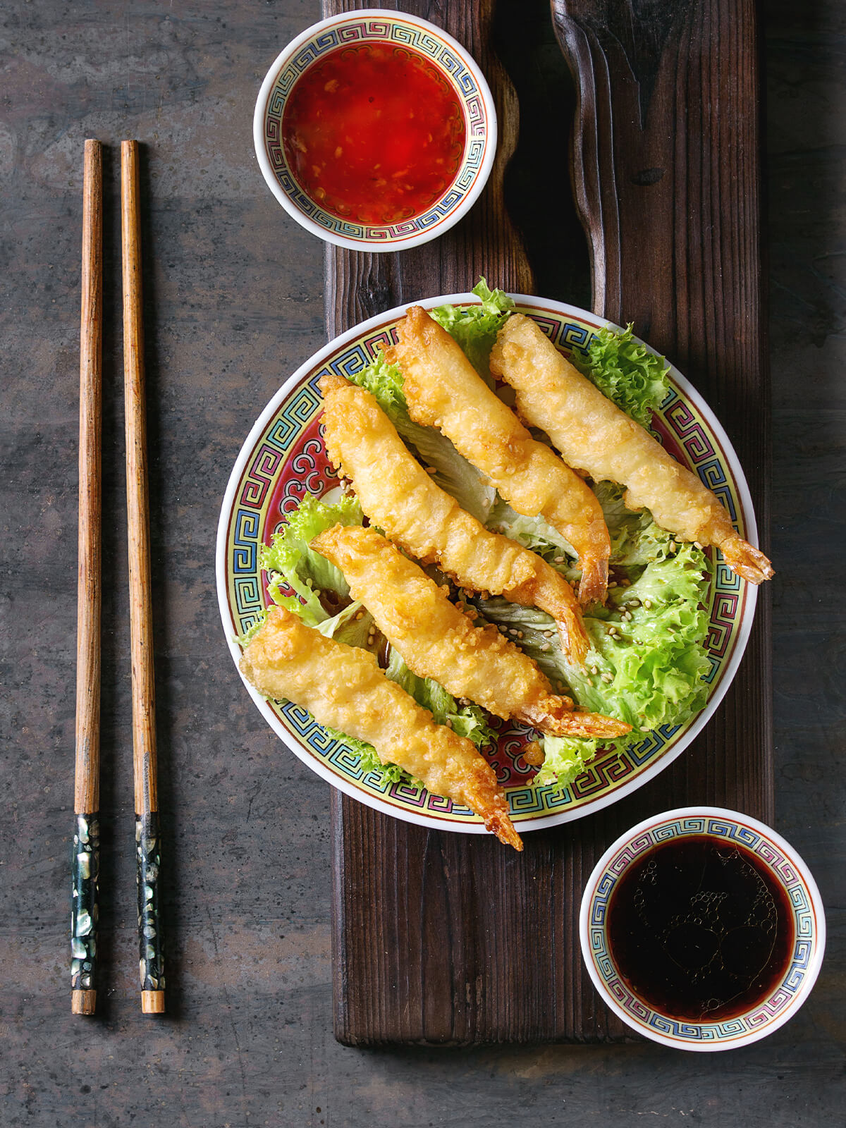 Fünf Tempura-Garnelen mit hell goldgelber, geschlossener Teighülle und teigfreien orange-bernsteinfarbenen Schwanzfächern, schräg überlappend auf krausem grünem Salat mit hellem Sesam, auf einem asiatischen Porzellanteller mit rotem Spiegel und buntem Mäanderrand; daneben ein Schälchen süße Chilisauce, ein Schälchen Sojasauce und Holzstäbchen, die links neben dem Holzbrett auf der dunklen Tischplatte liegen