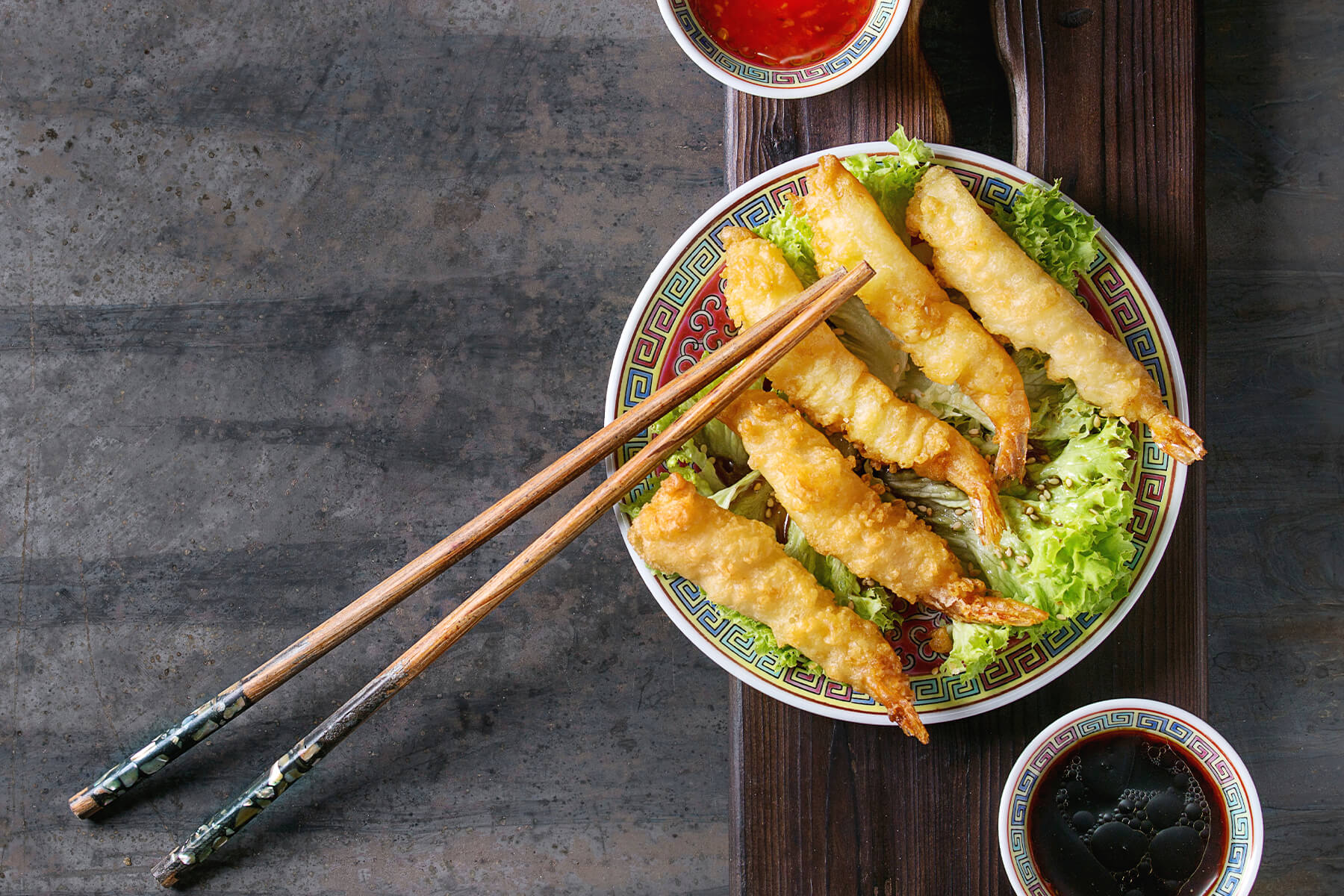 Tempura-Garnelen von oben auf einem asiatischen Porzellanteller mit rotem Spiegel und buntem Mäanderrand: fünf Garnelen mit hell goldgelber, geschlossener Teighülle und teigfreien Schwanzfächern, schräg nebeneinander auf krausem grünem Salat mit hellem Sesam, von links unten schräg über den Tellerrand bis etwa zur Tellermitte reichende Holzstäbchen, deren Perlmuttgriffe auf der dunklen Tischplatte liegen, dazu Schälchen mit süßer Chilisauce und Sojasauce