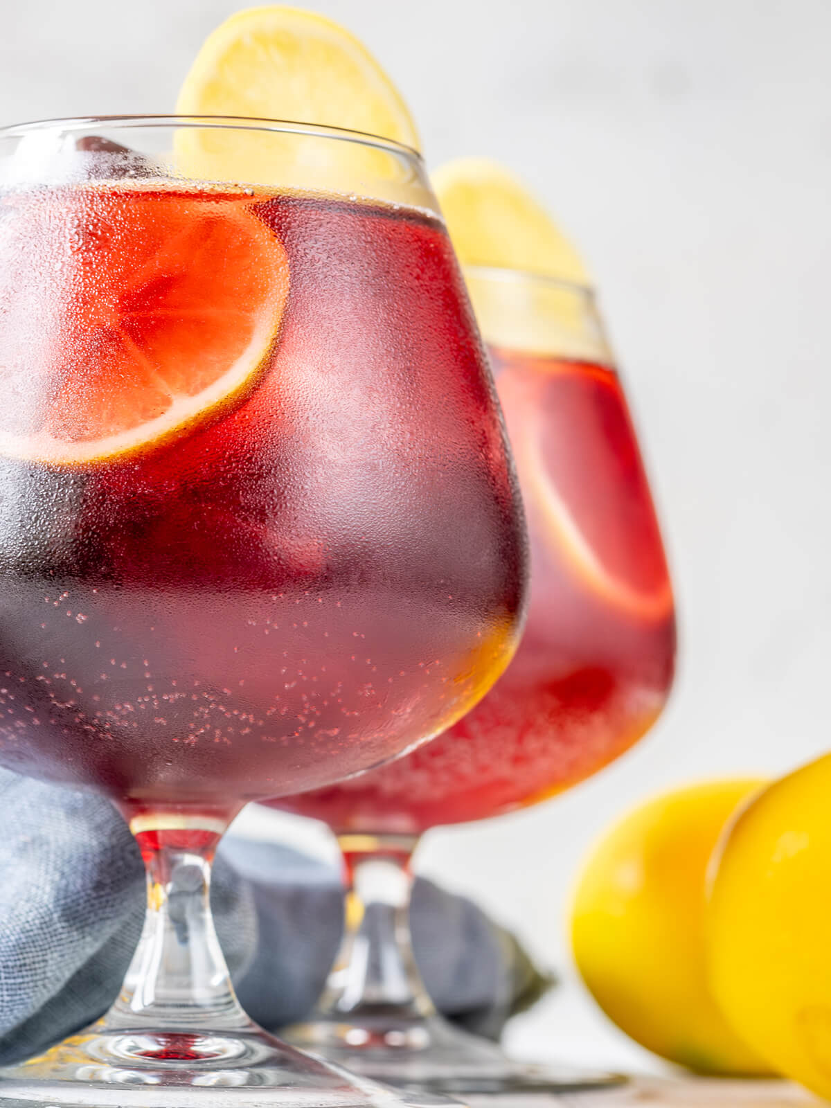 Nahaufnahme von rubinrotem Tinto de Verano im Stielglas mit feinen Bläschen, Eiswürfeln und einer Zitronenscheibe, daneben ganze Zitronen