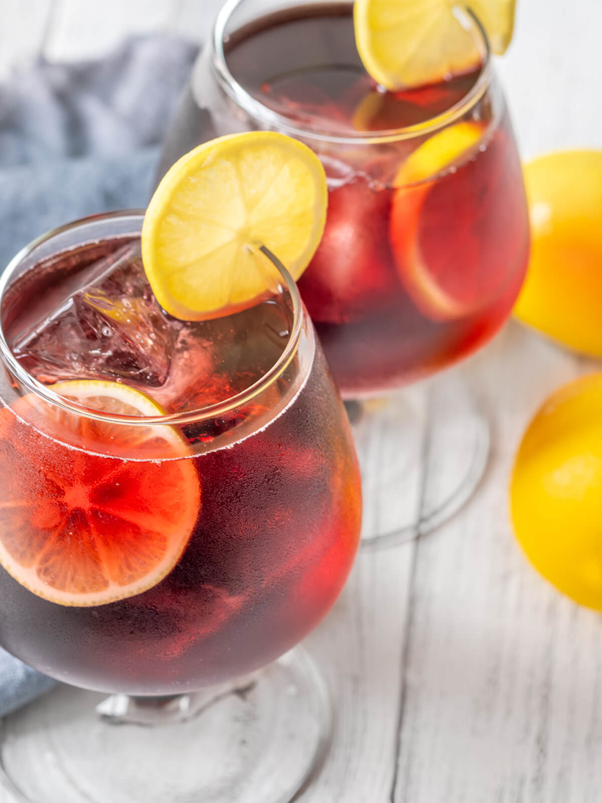 Zwei Stielgläser mit tief rubinrotem Tinto de Verano, gefüllt mit Eiswürfeln und Zitronenscheiben, daneben ganze Zitronen und Orangen auf hellem Holz