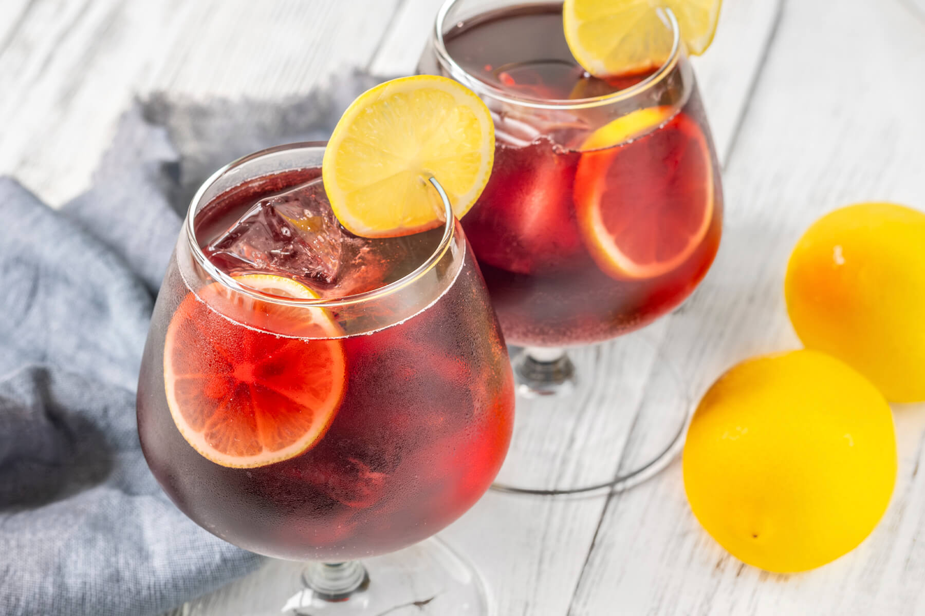 Zwei Gläser Tinto de Verano auf Eis mit schwimmenden Zitronenscheiben, daneben ganze Zitronen und Orangen auf hellem Holzuntergrund