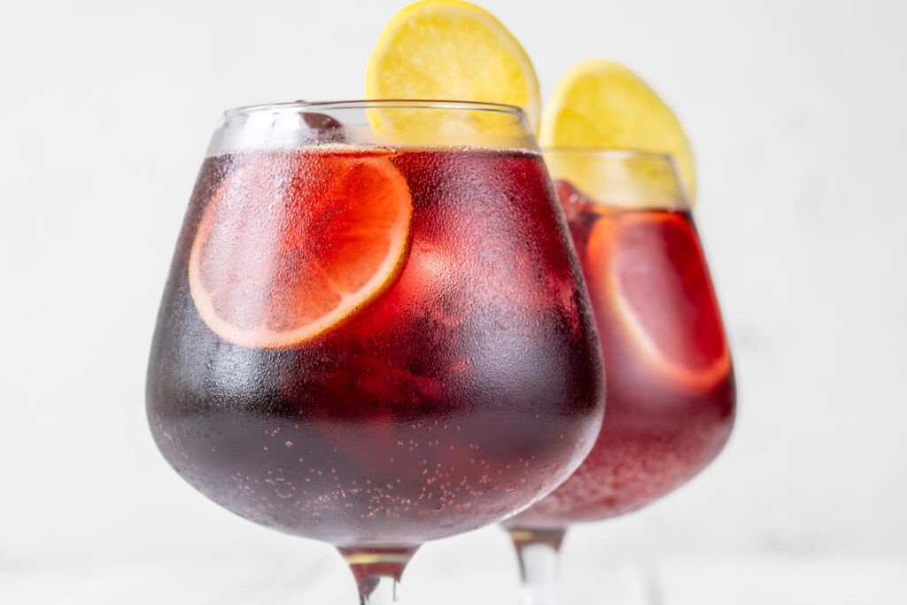 Zwei Stielgläser mit rubinrotem Tinto de Verano auf Eis, garniert mit Zitronen- und Orangenscheiben, feine Bläschen und beschlagene Gläser