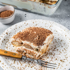 Ein Stück klassisches Tiramisu auf einem gesprenkelten Teller, mit Kakao bestäubt, die Löffelbiskuit- und Mascarponeschichten im Anschnitt sichtbar, dahinter die Glasform