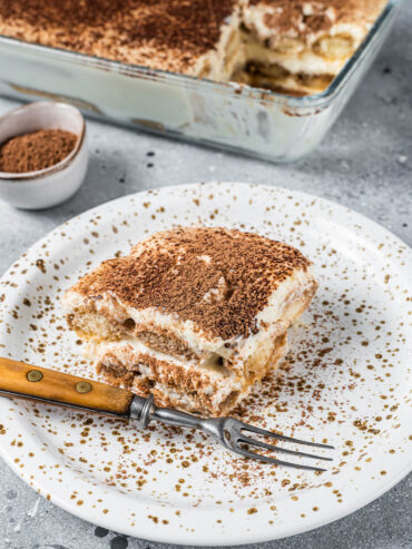 Ein Stück klassisches Tiramisu auf einem gesprenkelten Teller, mit Kakao bestäubt, die Löffelbiskuit- und Mascarponeschichten im Anschnitt sichtbar, dahinter die Glasform