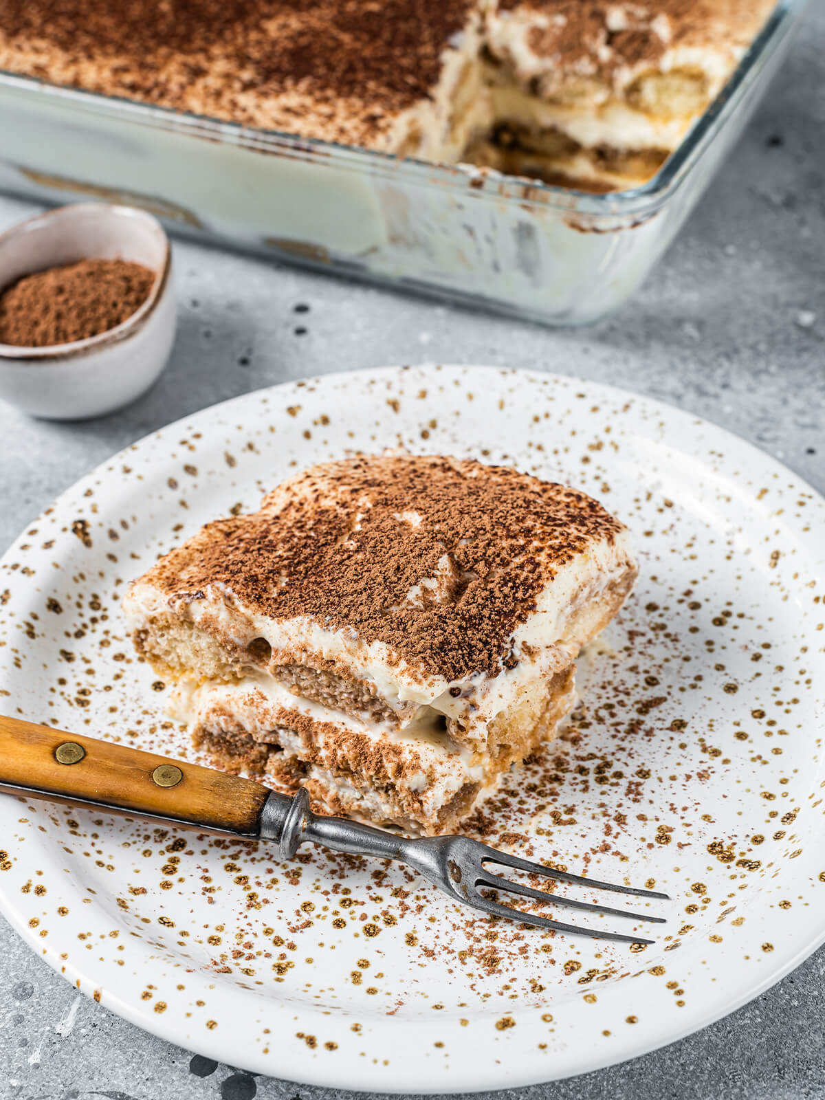 Ein Stück klassisches Tiramisu auf einem gesprenkelten Teller, mit Kakao bestäubt, die Löffelbiskuit- und Mascarponeschichten im Anschnitt sichtbar, dahinter die Glasform