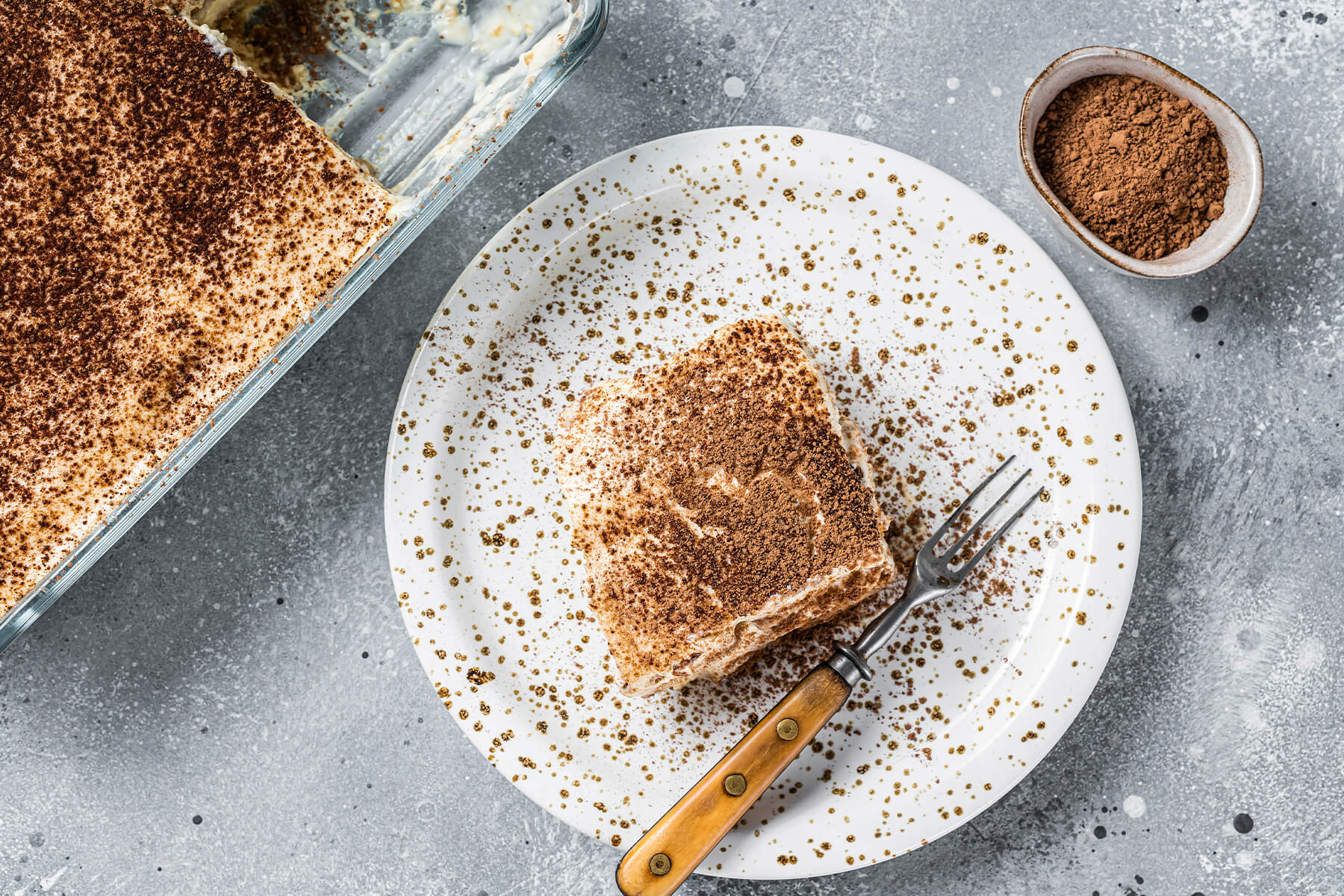 Draufsicht auf ein Stück Tiramisu mit Kakao auf einem gesprenkelten Teller, daneben die angeschnittene Glasform und ein Schälchen Kakao