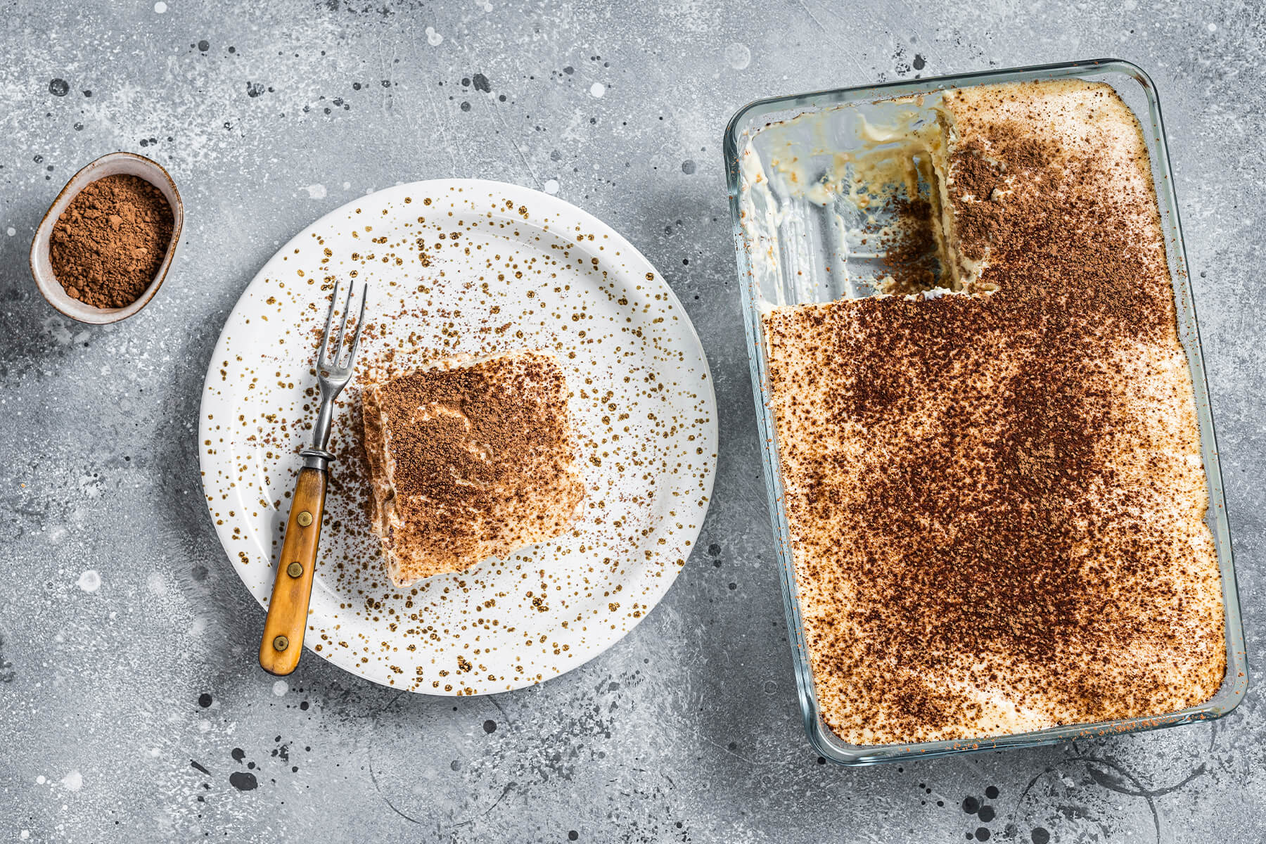 Draufsicht auf die angeschnittene rechteckige Glasform mit Kakaodecke, daneben ein Stück Tiramisu auf einem gesprenkelten Teller und ein Schälchen Kakao