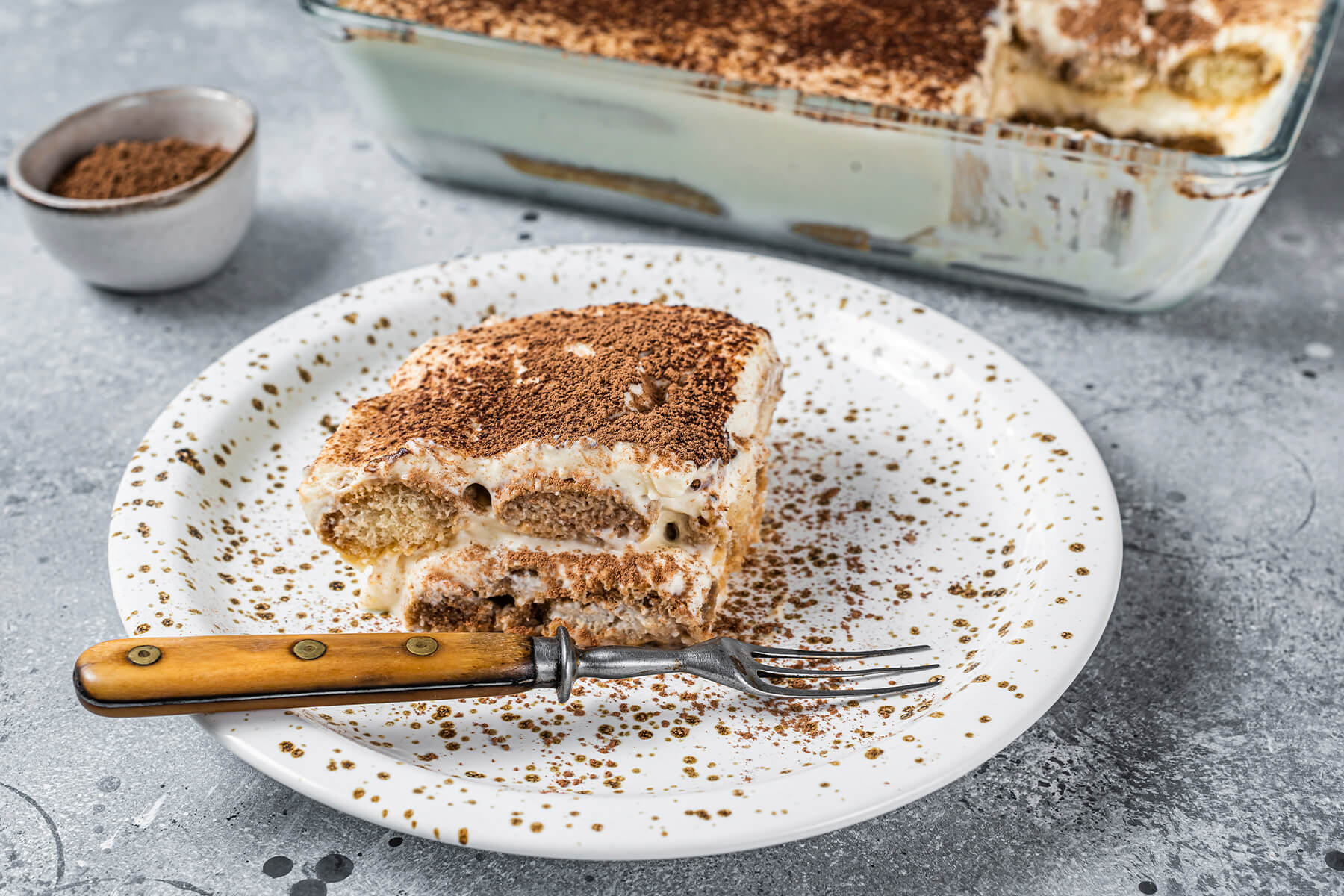 Ein Stück Tiramisu auf einem gesprenkelten Teller, im Hintergrund die Glasform, im Anschnitt zwei Lagen aus getränktem Biskuit und heller Creme