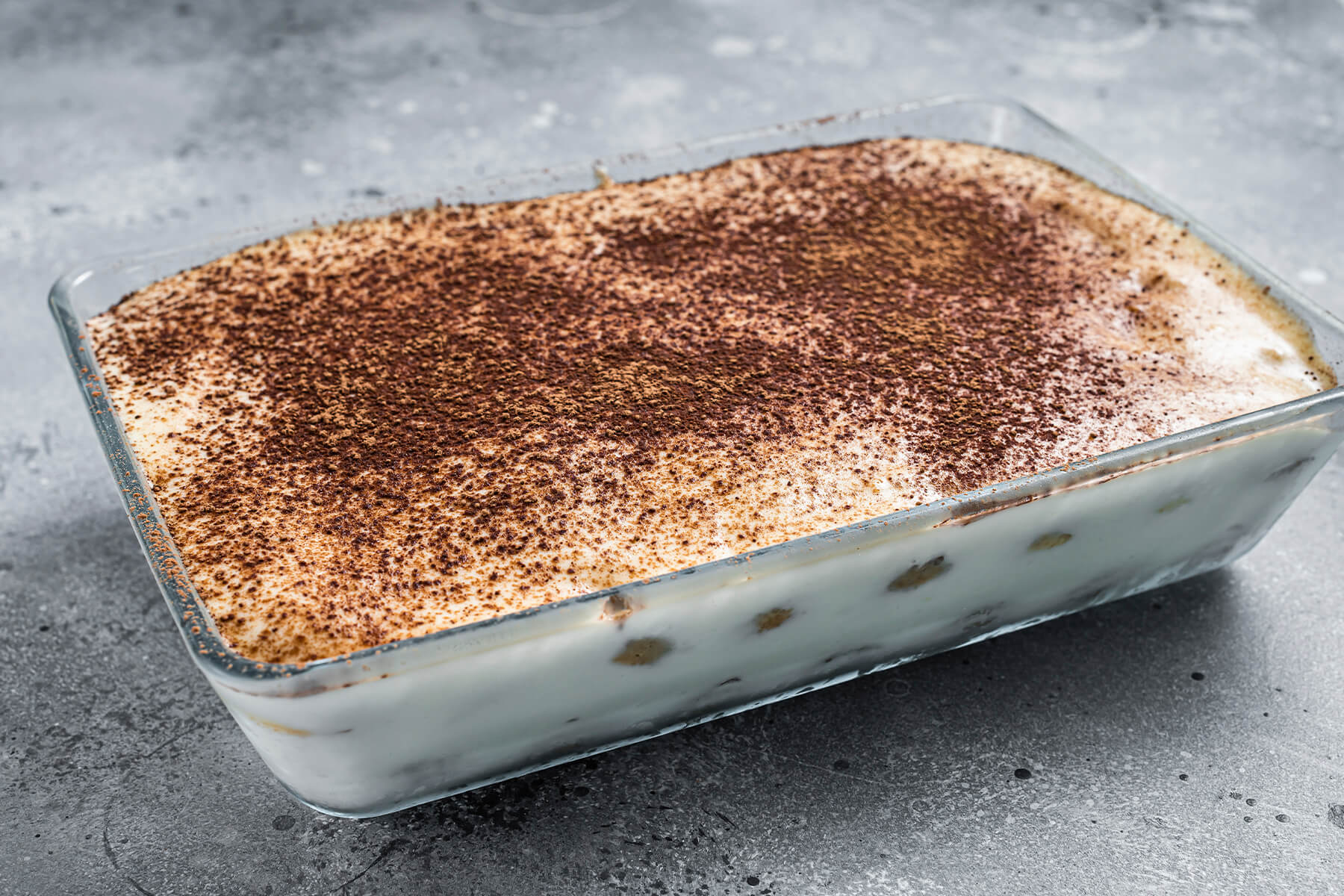 Die ganze mit Kakao bestäubte rechteckige Tiramisu-Form in Schrägansicht, an der Glaswand haftet Creme, dazwischen schimmern getränkte Biskuits durch