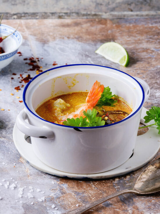 Tom Yam, scharf-saure Thai-Suppe mit ganzer Garnele, Shiitake und frischen Kräutern in cremig-oranger Brühe in einem weißen Topf, daneben Chiliflocken