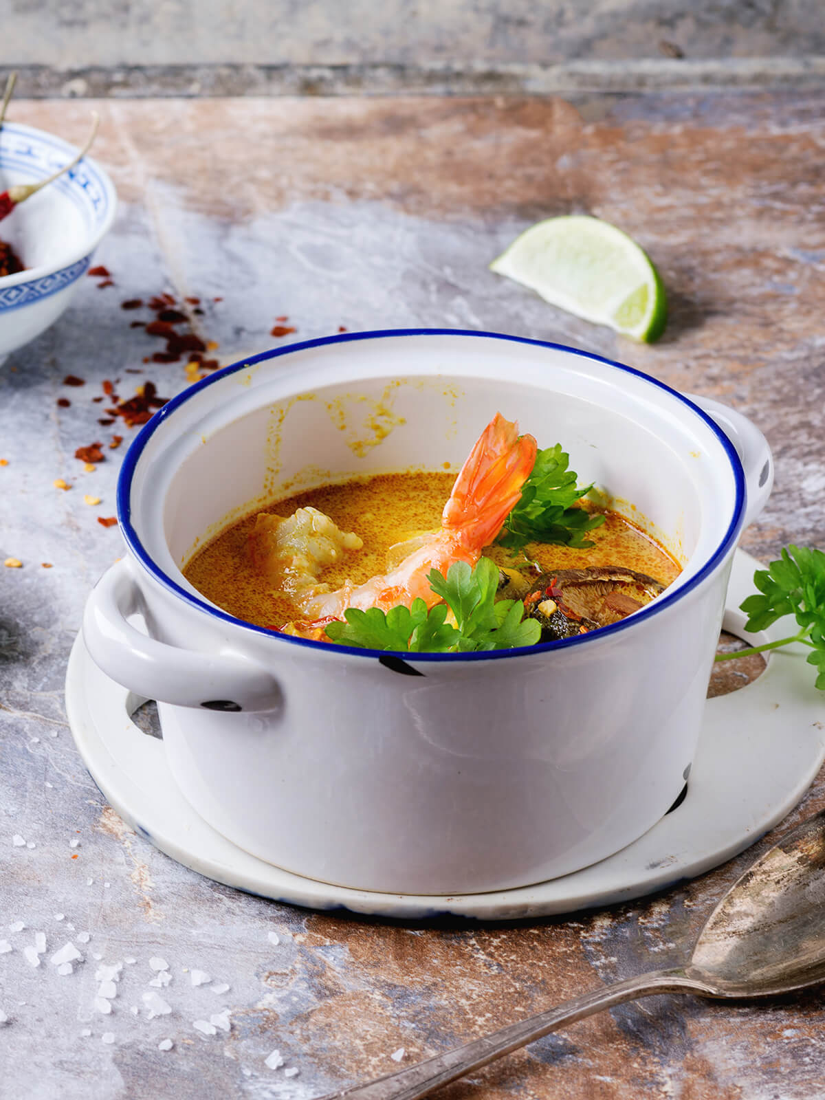 Tom Yam, scharf-saure Thai-Suppe mit ganzer Garnele, Shiitake und frischen Kräutern in cremig-oranger Brühe in einem weißen Topf, daneben Chiliflocken