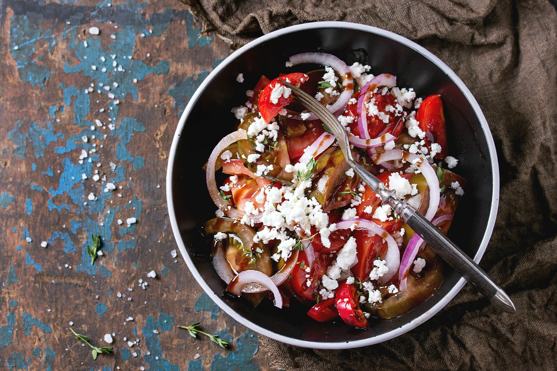 Blick von oben auf eine schwarze Schale mit schmalem weißem Rand auf dunklem Holz mit blau abgeblätterter Farbe: rote Tomatenspalten und dunkle grünlich-braune Tomatenscheiben mit rohen roten Zwiebelringen, grob zerkrümeltem weißem Feta und kleinen Thymianblättchen, dunkles Dressing am Boden; eine alte Gabel liegt im Salat, rechts ein braunes Jutetuch, links grobe Salzkörner verstreut