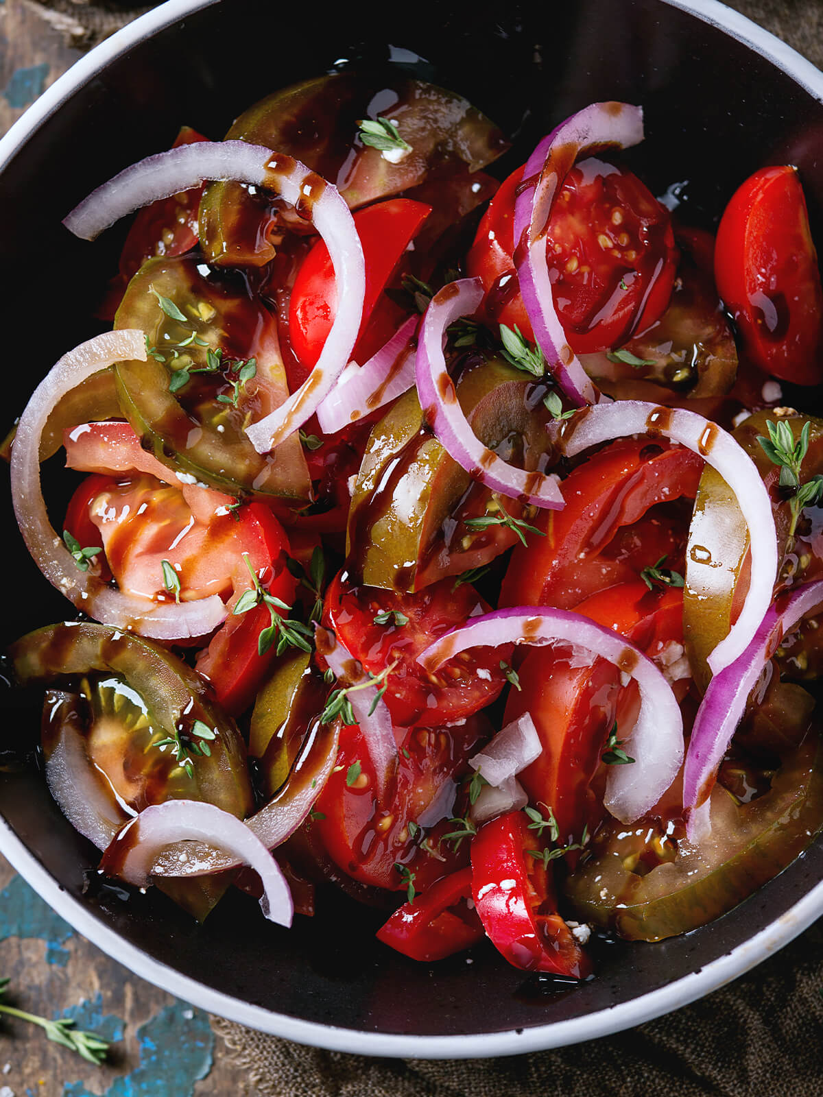 Nahaufnahme in einer schwarzen Schale: rote Tomatenspalten und dunkle grünlich-braune Tomatenscheiben, darüber rohe rote Zwiebelringe und feine Thymianblättchen, alles glänzend von dunklem Öl-Balsamico-Dressing, das sich am Schalenboden sammelt; nur vereinzelt kleine weiße Fetakrümel dazwischen