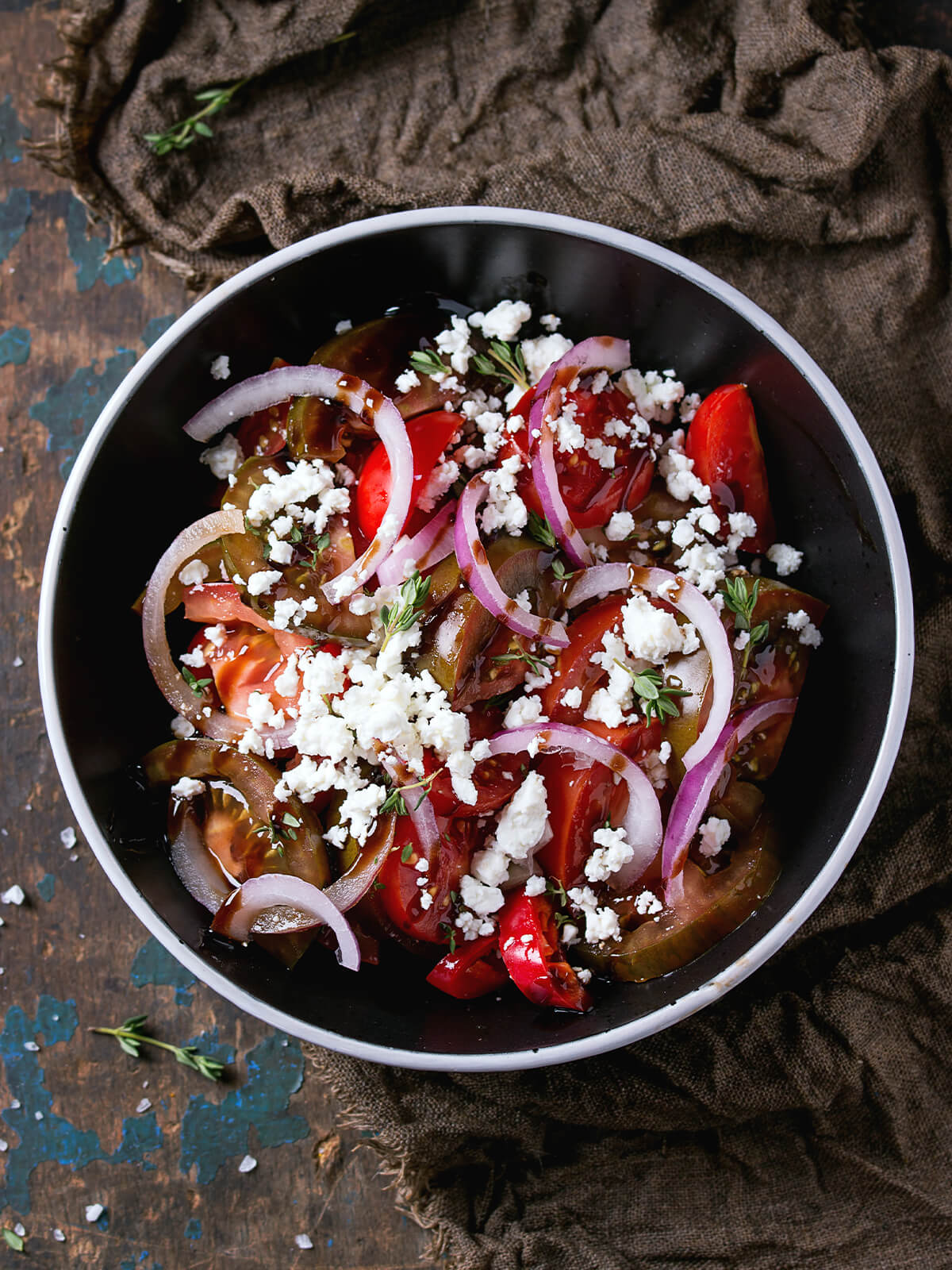 Blick von oben auf eine schwarze Schale mit schmalem weißem Rand, gefüllt mit roten Tomatenspalten und dunklen grünlich-braunen Tomatenscheiben, rohen roten Zwiebelringen, grob zerkrümeltem weißem Feta und kleinen Thymianblättchen; dunkles Öl-Balsamico-Dressing sammelt sich am Schalenboden, die Schale steht auf einem braunen Jutetuch neben dunklem Holz mit blau abgeblätterter Farbe