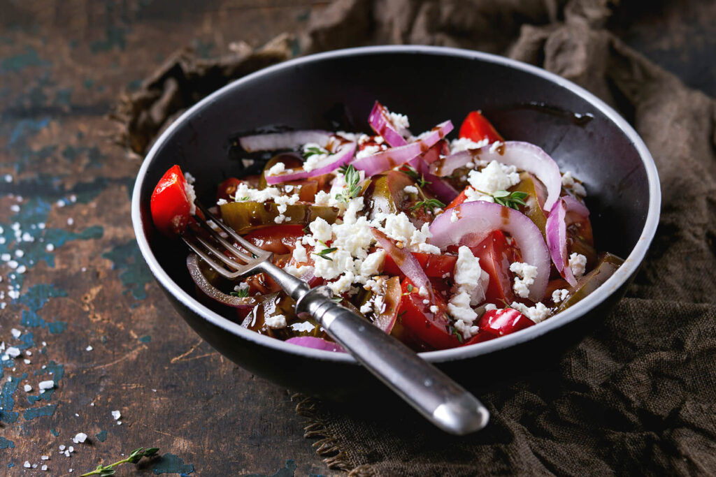 Schräg von der Seite fotografierte schwarze Schale mit schmalem weißem Rand: bunter Tomatensalat aus roten Spalten und dunklen grünlich-braunen Scheiben mit rohen roten Zwiebelringen, grob zerkrümeltem weißem Feta und Thymianblättchen, dunkles Dressing glänzt zwischen den Stücken; eine alte Gabel steckt im Salat, links grobe Salzkörner auf dunklem Holz mit blau abgeblätterter Farbe, rechts ein braunes Jutetuch
