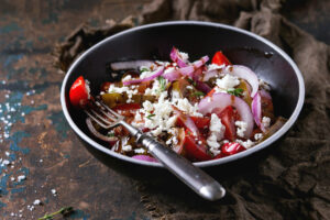 Schräg von der Seite fotografierte schwarze Schale mit schmalem weißem Rand: bunter Tomatensalat aus roten Spalten und dunklen grünlich-braunen Scheiben mit rohen roten Zwiebelringen, grob zerkrümeltem weißem Feta und Thymianblättchen, dunkles Dressing glänzt zwischen den Stücken; eine alte Gabel steckt im Salat, links grobe Salzkörner auf dunklem Holz mit blau abgeblätterter Farbe, rechts ein braunes Jutetuch