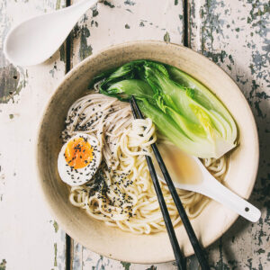 Japanische Udon-Suppe mit dicken Nudeln, Pak Choi, Enoki, weichem Ei und schwarzem Sesam in einer hellen Schüssel