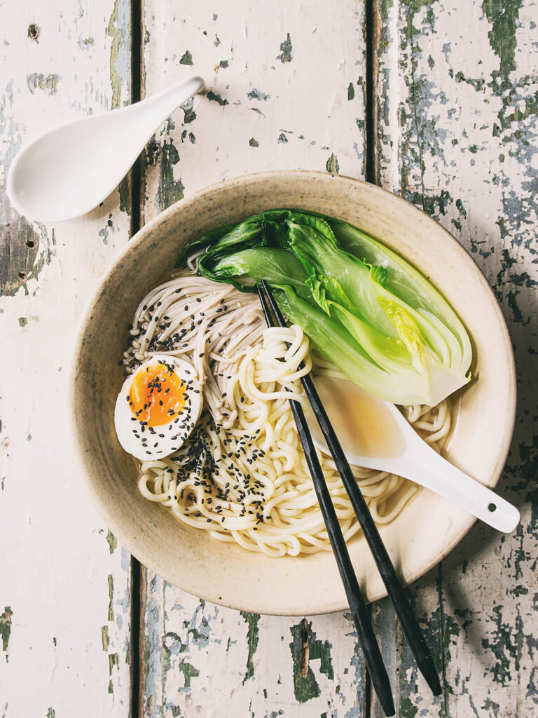 Japanische Udon-Suppe mit dicken Nudeln, Pak Choi, Enoki, weichem Ei und schwarzem Sesam in einer hellen Schüssel