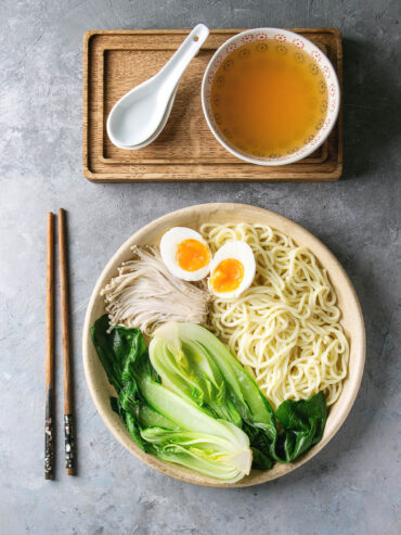 Japanische Udon-Suppe mit dicken Nudeln, Pak Choi, Enoki und weichem Ei auf einem Teller, daneben ein Schälchen klare Brühe auf einem Holzbrett