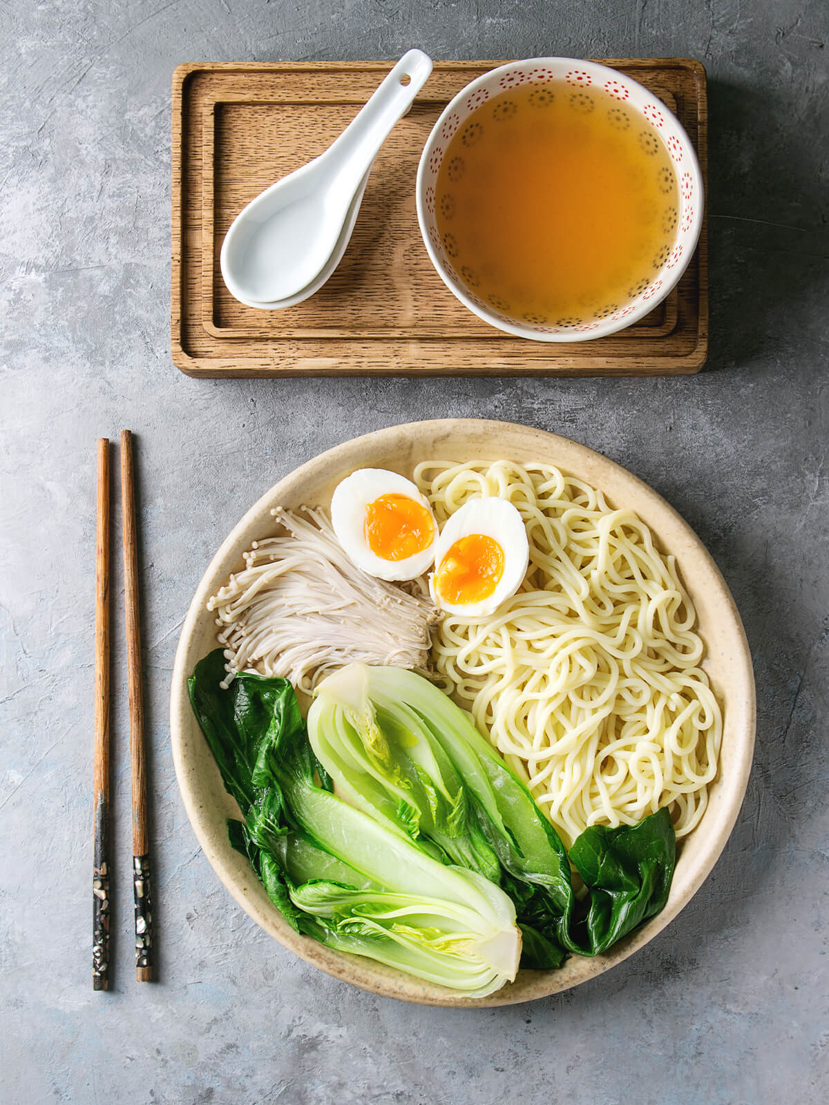 Japanische Udon-Suppe mit dicken Nudeln, Pak Choi, Enoki und weichem Ei auf einem Teller, daneben ein Schälchen klare Brühe auf einem Holzbrett