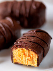 Angeschnittener dunkel glasierter Riegel auf einem weißen Teller, im Anschnitt die warm gelbe, körnige Cashew-Aprikosen-Masse mit deutlich sichtbaren orangen Aprikosenstückchen, die dicke Hülle mit einer erhabenen Schokoladen-Linie verziert