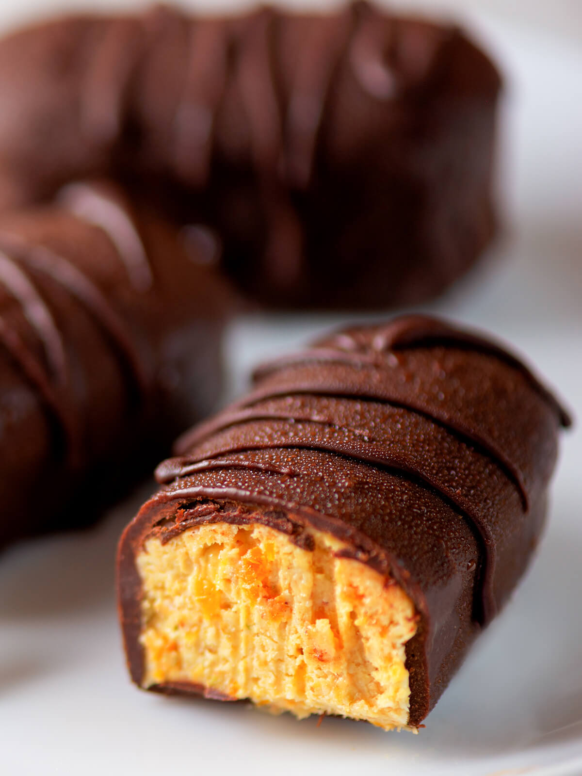 Angeschnittener dunkel glasierter Riegel auf einem weißen Teller, im Anschnitt die warm gelbe, körnige Cashew-Aprikosen-Masse mit deutlich sichtbaren orangen Aprikosenstückchen, die dicke Hülle mit einer erhabenen Schokoladen-Linie verziert