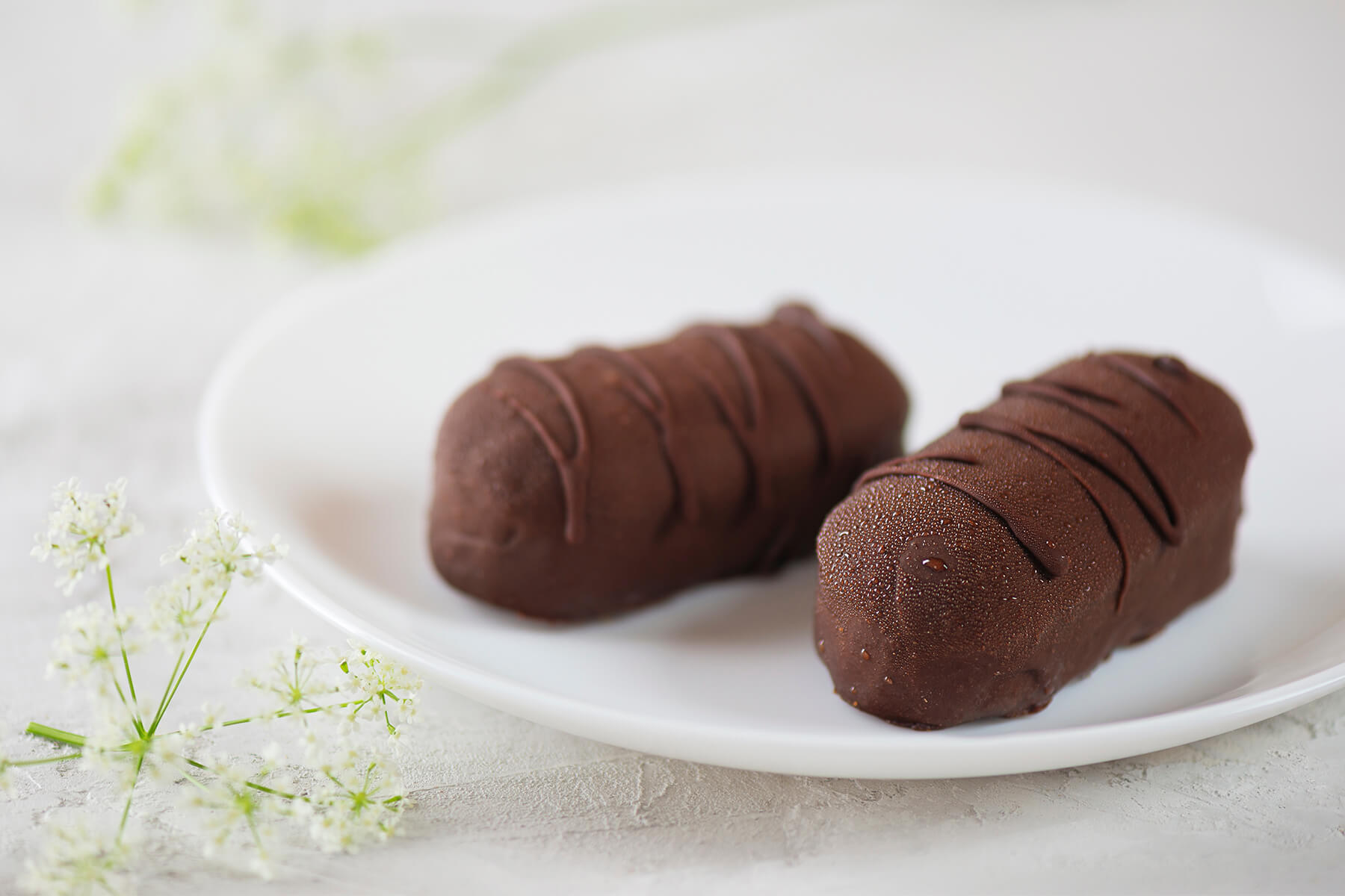 Zwei kräftige, mit dunkler Schokolade glasierte Cashew-Aprikosen-Riegel auf einem runden weißen Teller, jeweils mit erhabenen Schokoladen-Linien im Zickzack verziert, daneben weiße Doldenblüten als reine Deko